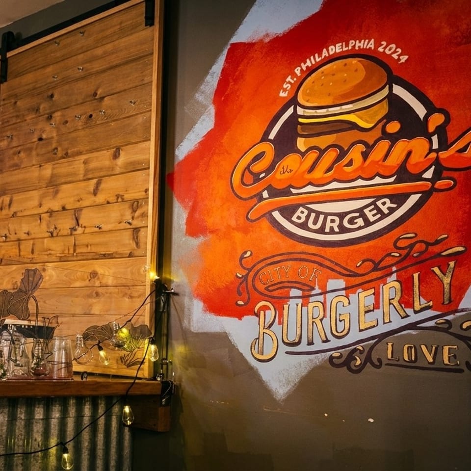 Cousin's Burger mural on a dark wall, with a wooden sliding door and string lights nearby.
