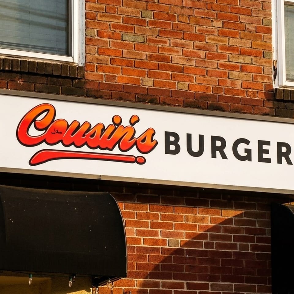 Cousin's Burger sign on a brick building with two windows and black awnings.