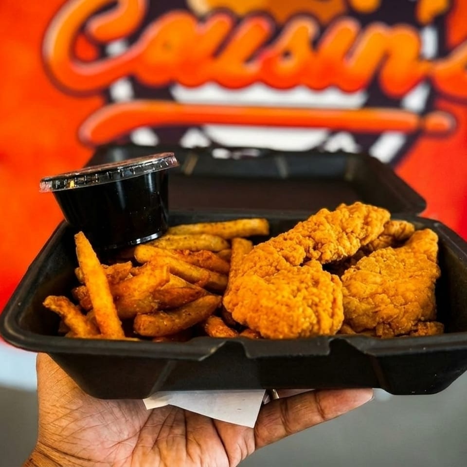 Hand holding a takeout container with crispy fried chicken tenders, seasoned fries, and dipping sauce.