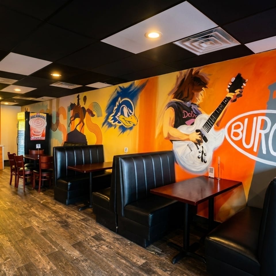 A restaurant interior with a person at the counter, booths, tables, and colorful murals on the walls.