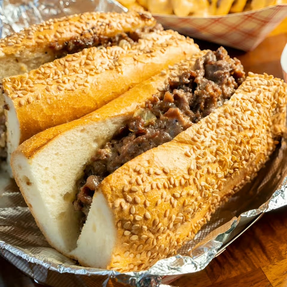 Steak and cheese sandwich on a sesame-seed roll with cheese fries in the background.